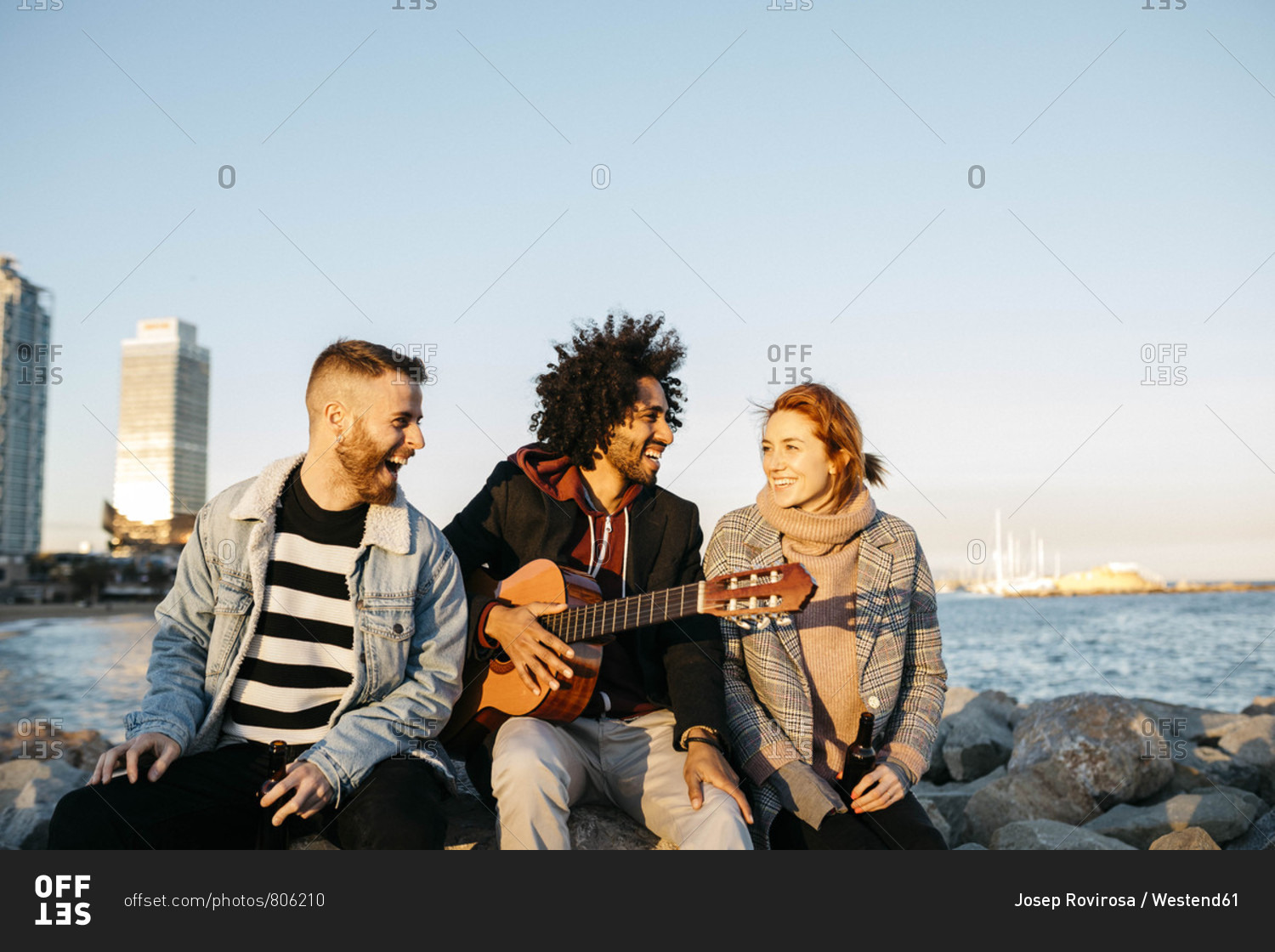 Three happy friends with guitar sitting outdoors at the coast sunset