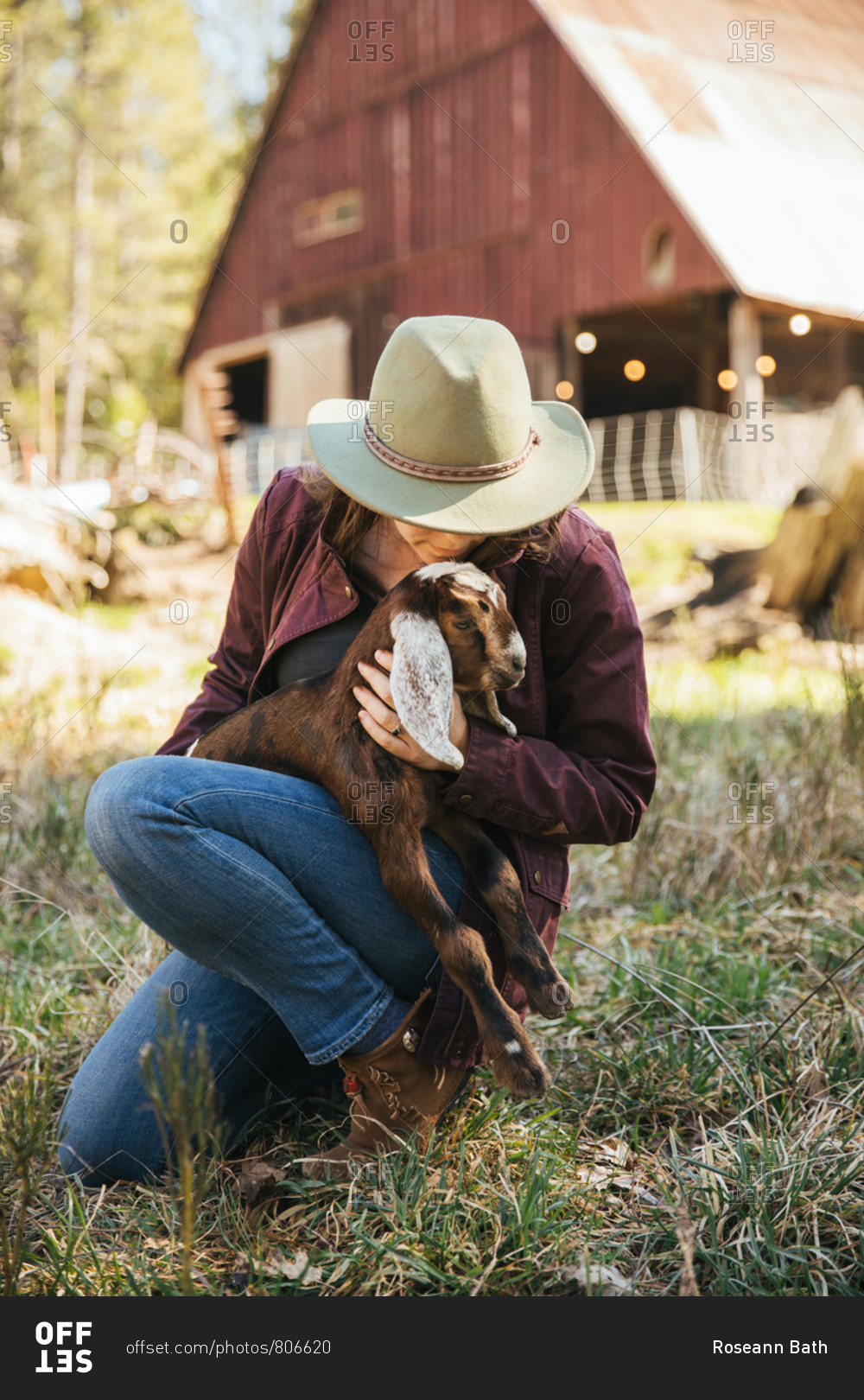 Woman cuddling a baby goat in front of a barn.