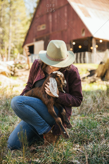 Woman cuddling a baby goat in front of a barn.