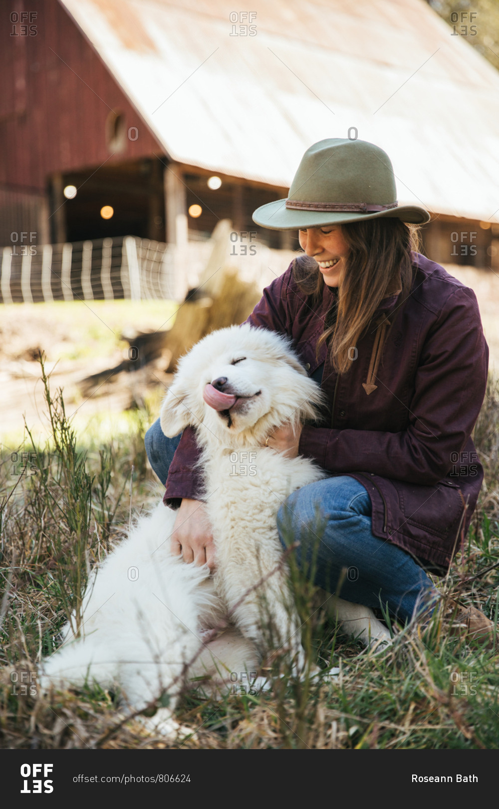 Woman cuddling a great pyrenees puppy in front of a barn while puppy licks lips.