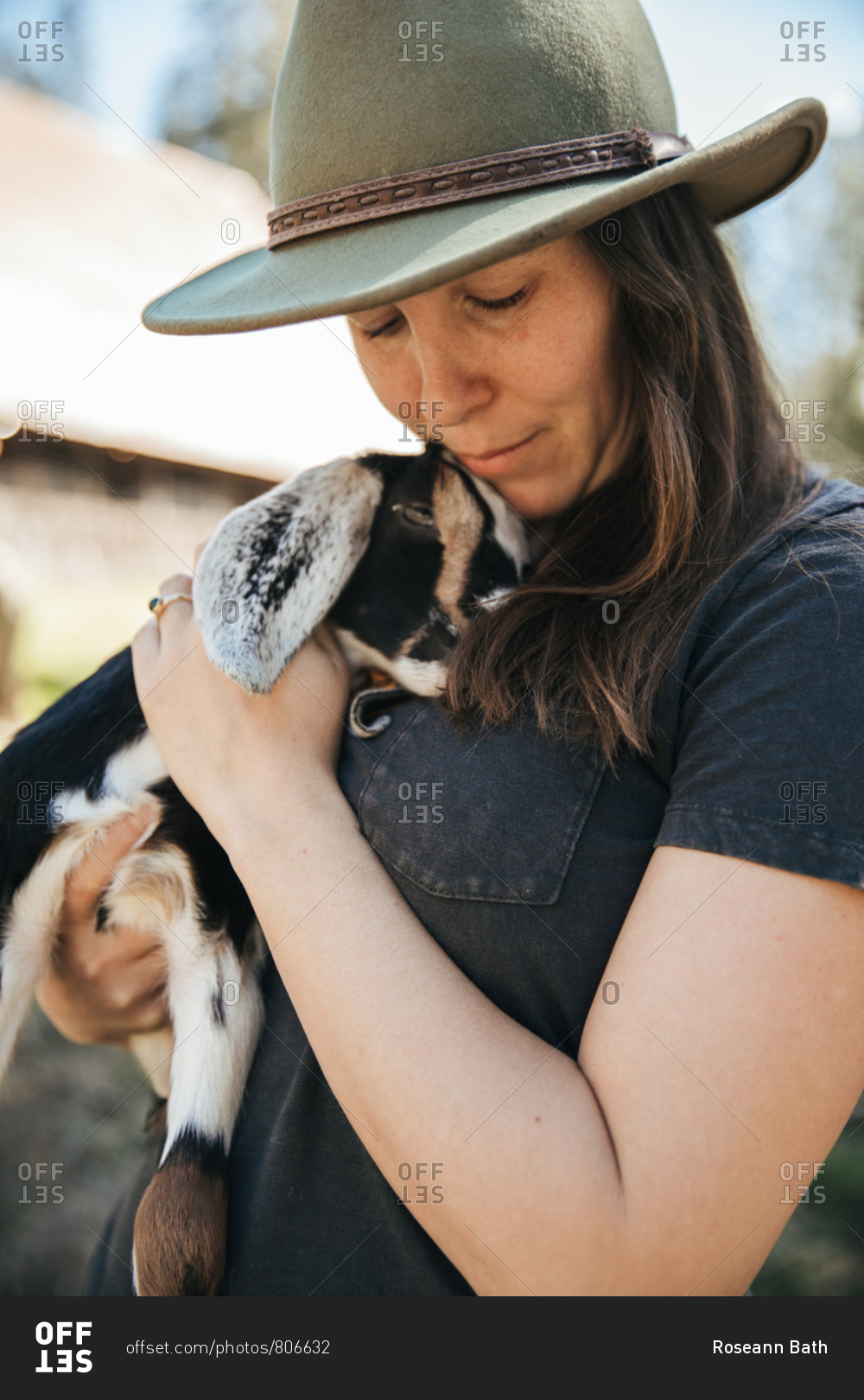 Woman in a hat holding a baby goat in front of  a barn.