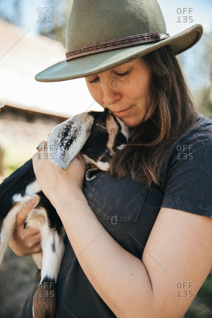 Woman in a hat holding a baby goat in front of  a barn.