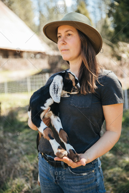 Woman in a hat holding a baby goat in front of  a barn.