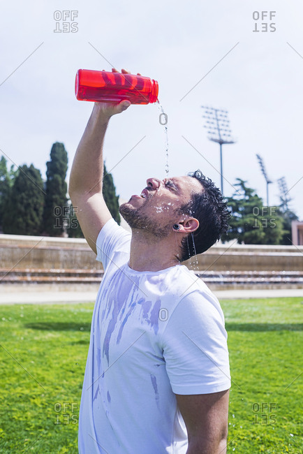 Side view of man pouring water from bottle on head in sportswear in park near fountain in sunny day