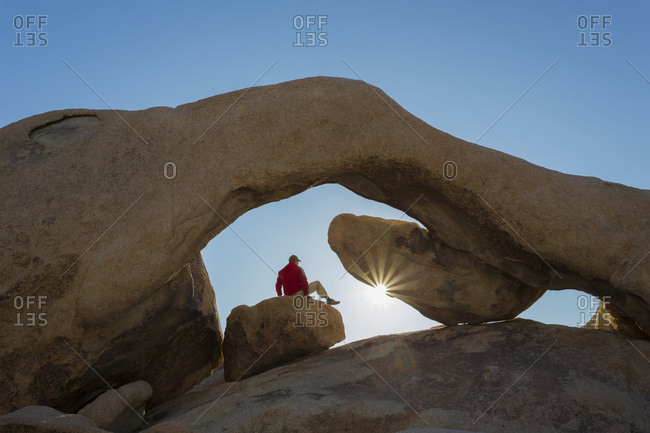 A hiker watching a sunrise under sandstone rock arch with sun star, Joshua Tree National Park; California, United States of America