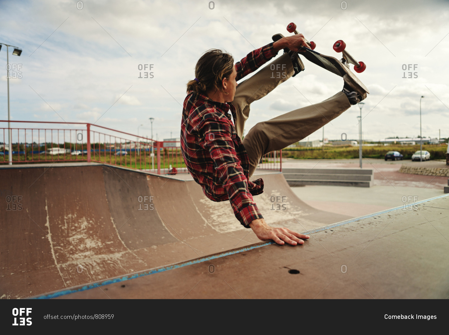 Side view skilled young skateboarder in checkered shirt doing handstand trick skate park under cloudy sky stock photo - OFFSET