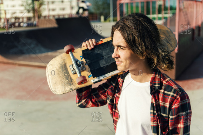 Portrait of handsome young with long hair looking away while carrying skateboard on shoulder in skate park stock photo - OFFSET