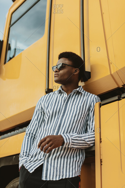 Portrait of a stylish black man wearing blue striped collared shirt leaning on a large yellow vehicle