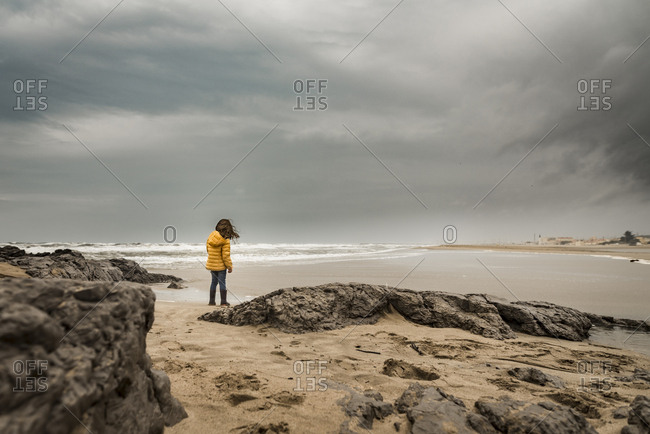 Girl struggles to walk along rocky beach in storm with cloudy sky