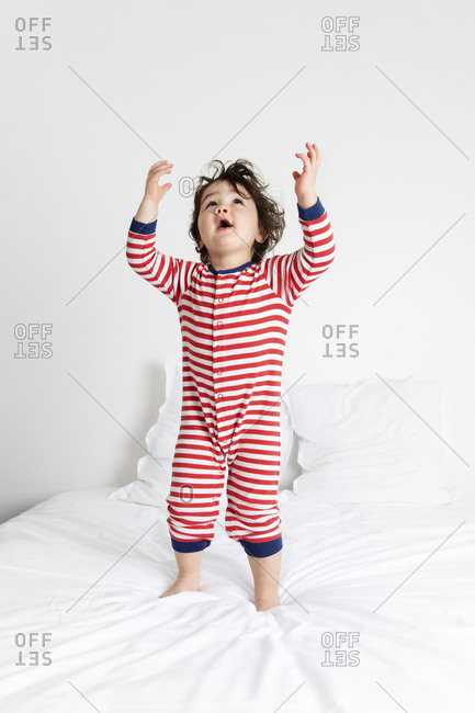 Cute little boy standing on white bed looking up with arms raised