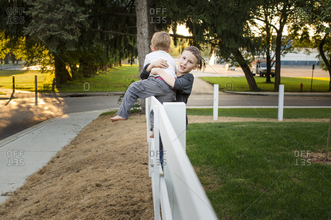 A boy helps his little brother get down from a fence