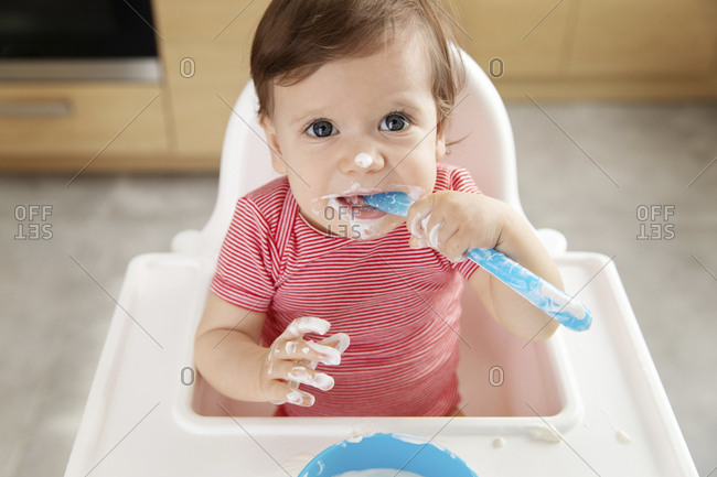 Messy baby in high chair eating yogurt with spoon
