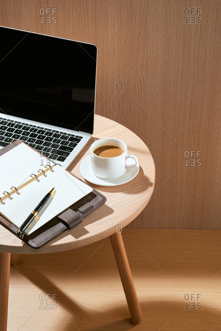 Wood office desk table with blank notebook, laptop computer, cup of coffee and supplies.