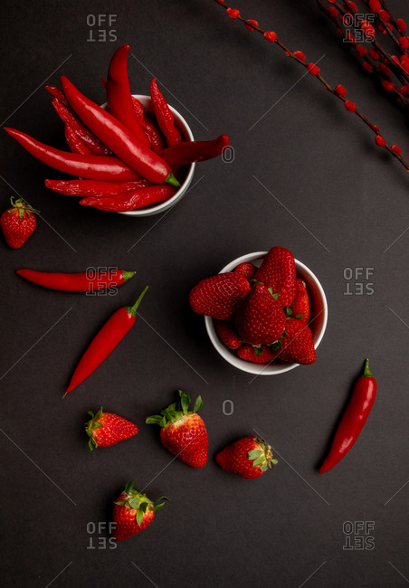 Red fabric and twigs with bright buds placed on black background near hot chili peppers and sweet ripe strawberries