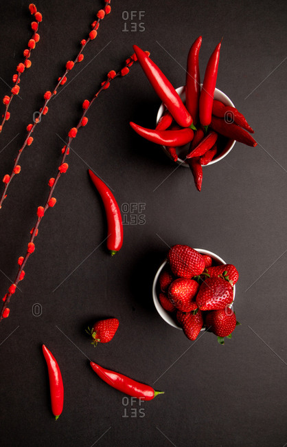 Red fabric and twigs with bright buds placed on black background near hot chili peppers and sweet ripe strawberries