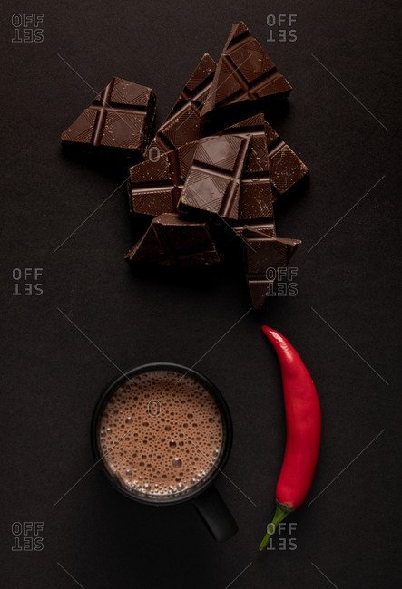 Pieces of yummy chocolate placed near black mug of steamy hot beverage with chili pepper on dark background