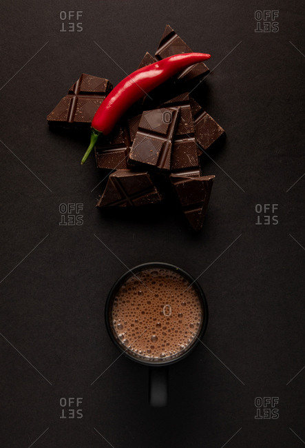 Pieces of yummy chocolate placed near black mug of steamy hot beverage with chili pepper on dark background
