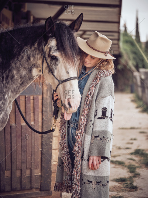 Side view of attractive cowgirl with closed eyes touching head of dapple gray horse while standing near stable on blurred background of ranch