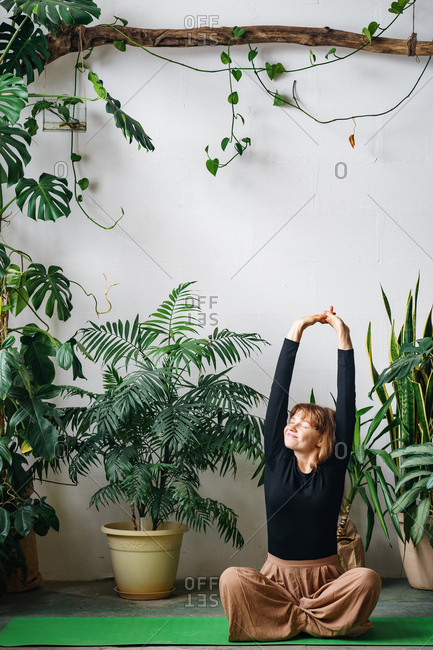 Redheaded young woman stretching sitting on a green yoga mat surrounded by lots of house plants