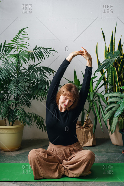 Redheaded young woman stretching sitting on a green yoga mat surrounded by lots of home plants