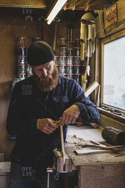 Bearded man wearing black beanie standing at workbench in workshop, working on piece of wood.
