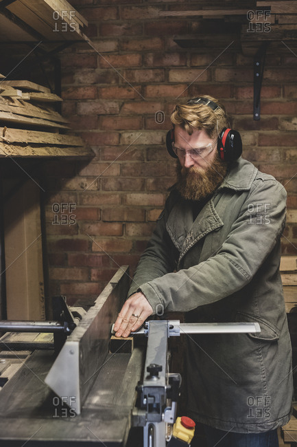 Bearded man standing in workshop, wearing ear protectors, working on piece of wood.