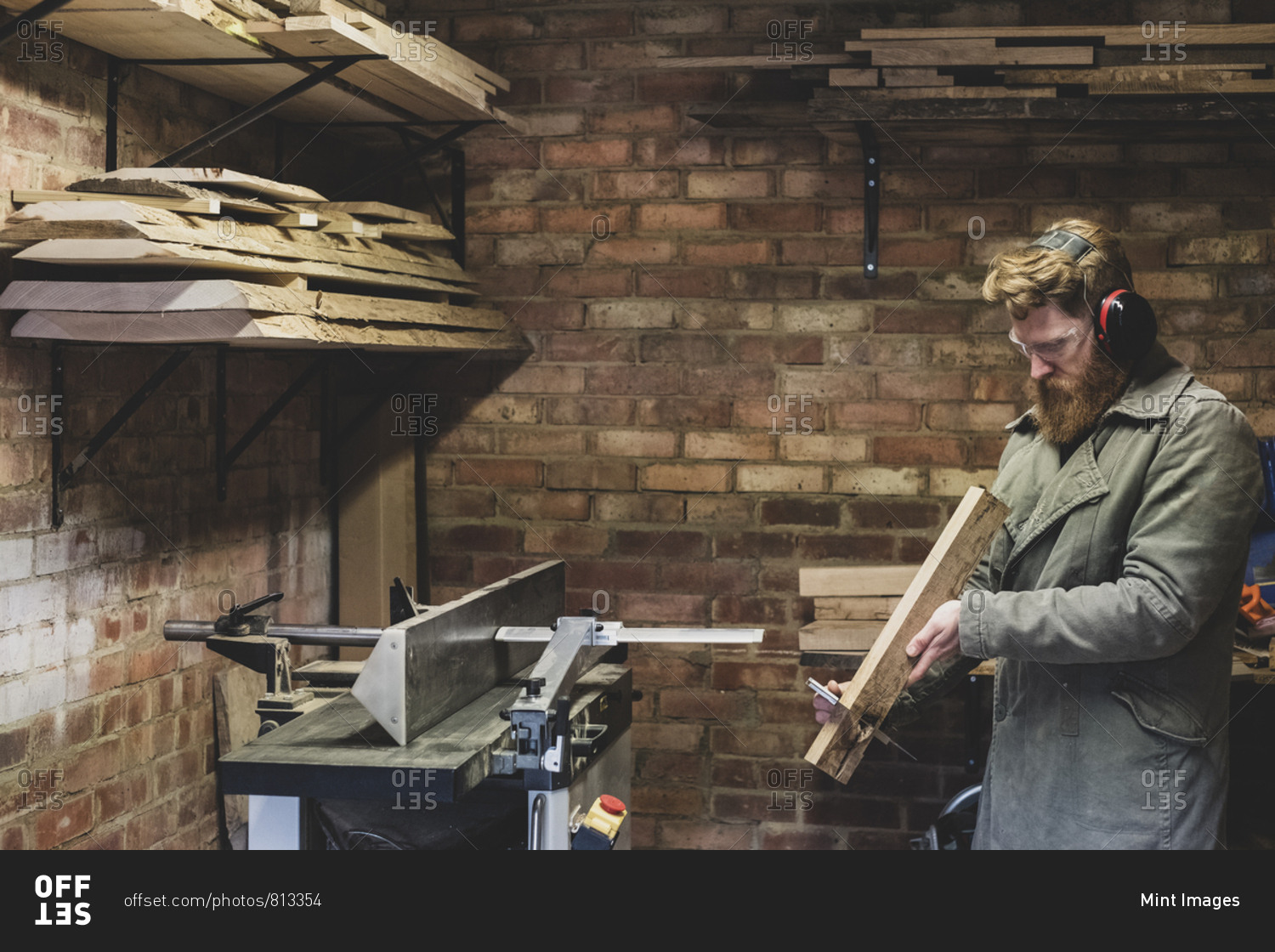 Bearded man standing in workshop, wearing ear protectors, working on piece of wood.