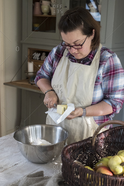 Woman wearing apron standing in a kitchen cutting butter pieces into a metal bowl filled with flour.