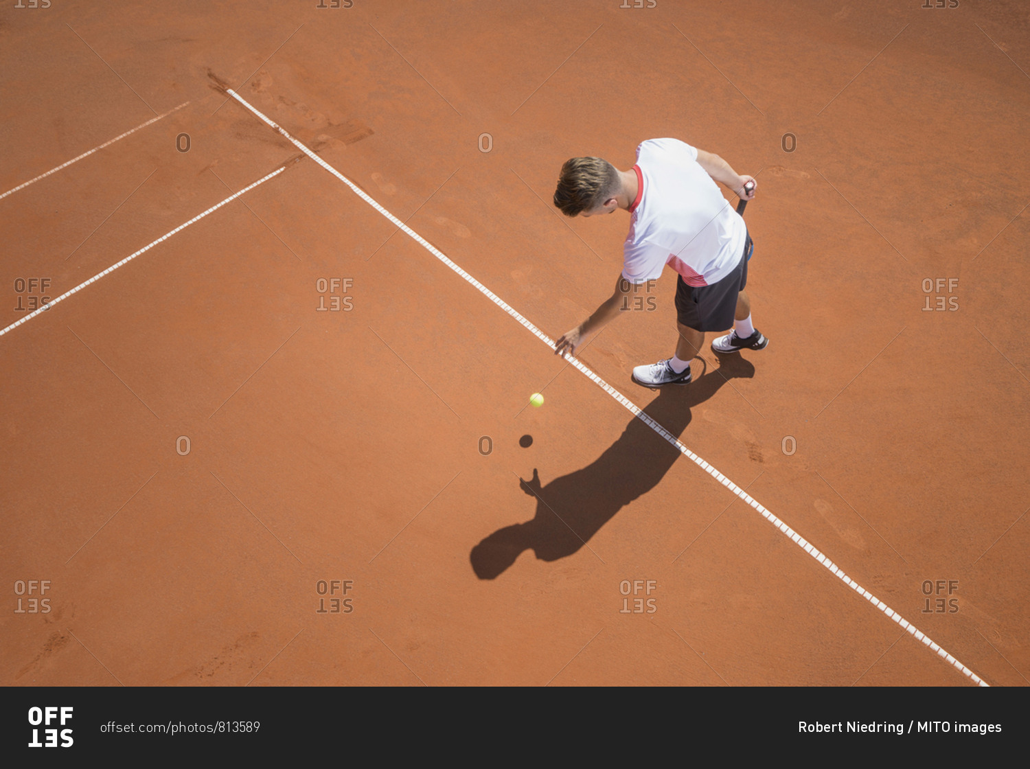 Young male tennis player preparing to serve the ball on sunny red tennis court, Bavaria, Germany