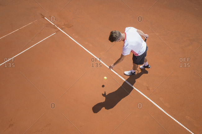 Young male tennis player preparing to serve the ball on sunny red tennis court, Bavaria, Germany