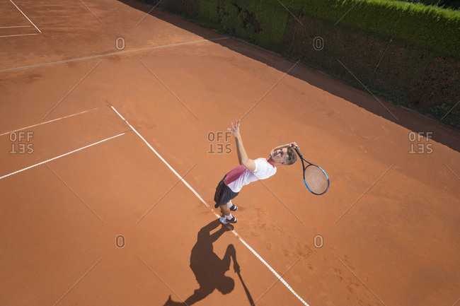 Young male tennis player serving the ball on sunny red tennis court, Bavaria, Germany