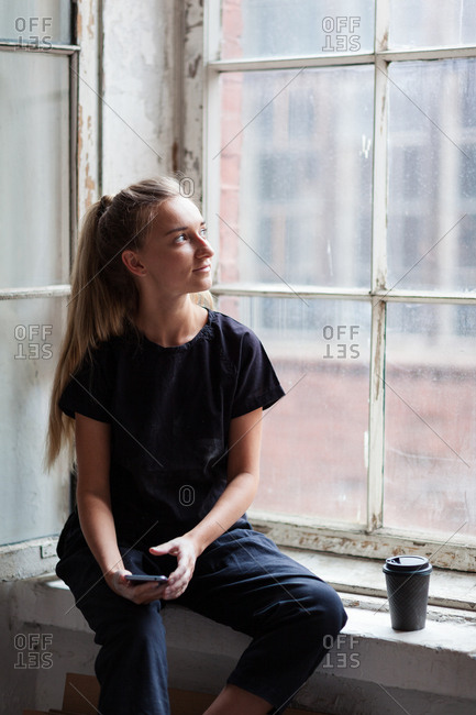 Creative young woman sitting on window sill with cell phone in her hand and looking through old dirty glass dreamily, paper cup of coffee placed nearby