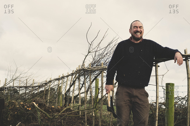 Bearded man holding axe and bill hook standing next to a newly built traditional hedge, laughing.