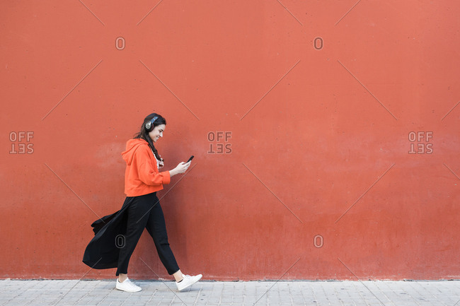 Young dancer walking and using the phone and headphones in front of a red wall