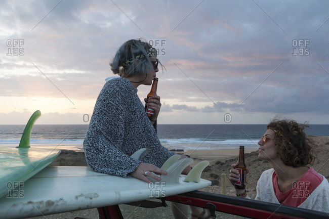 Two surfer girls with sunglasses on their 4x4 car drinking beer at sunset
