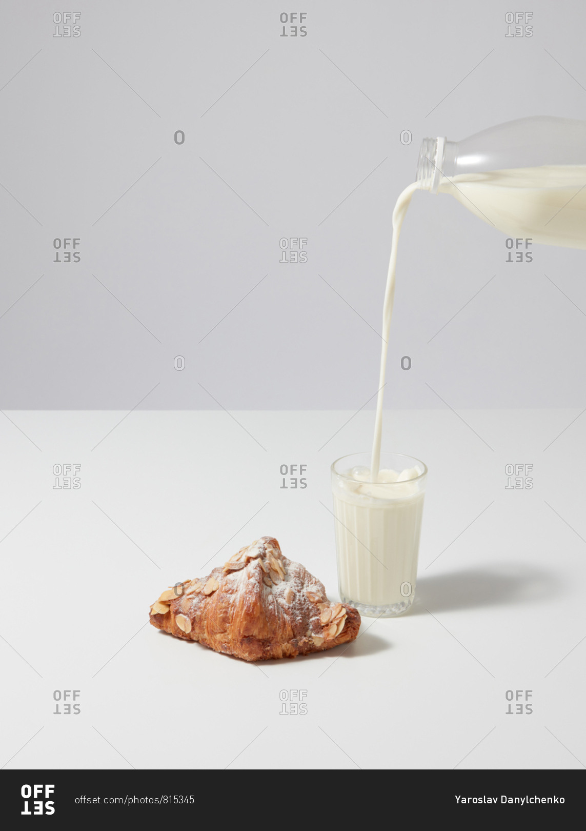 Fresh milk is pouring into a glass from bottle and fresh homemade croissant with powdered sugar and almond chips on a gray background, copy space. Continental morning breakfast.