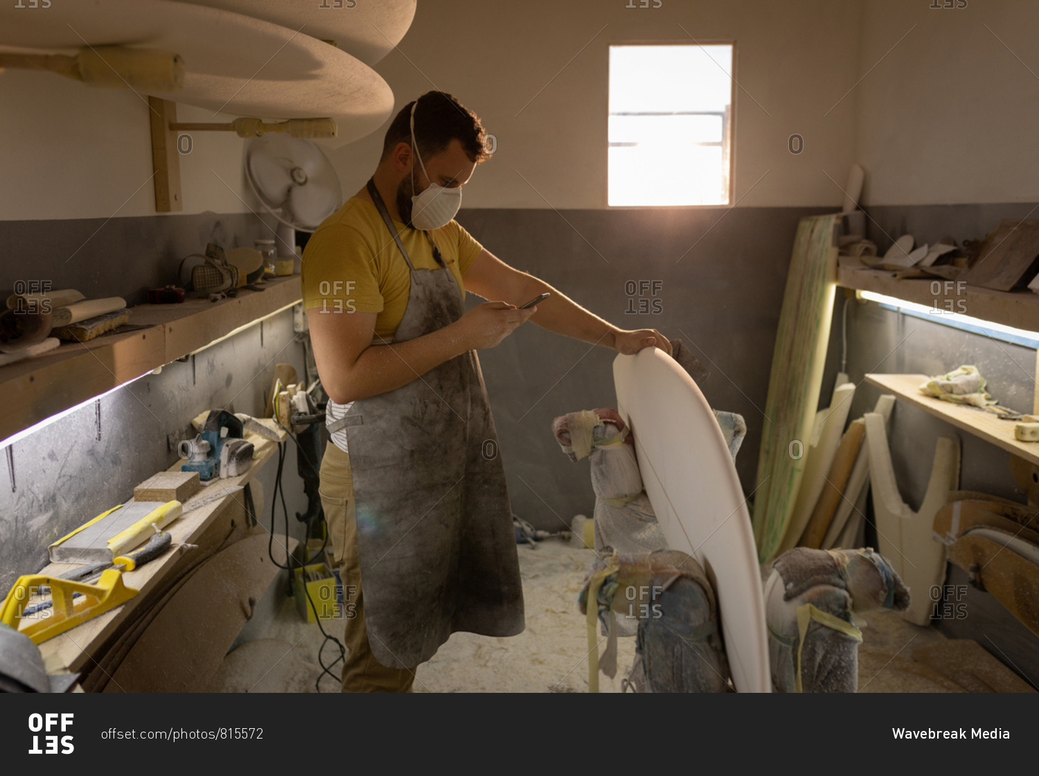 Side view of Caucasian man with mouth protection mask using mobile phone while cleaning surfboard in a workshop
