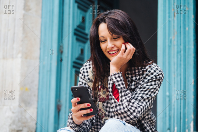 Young Brunette Woman With Red And Plaid Clothes Sitting In Front Of An Old Building With A Blueish Door While Using Her Phone.