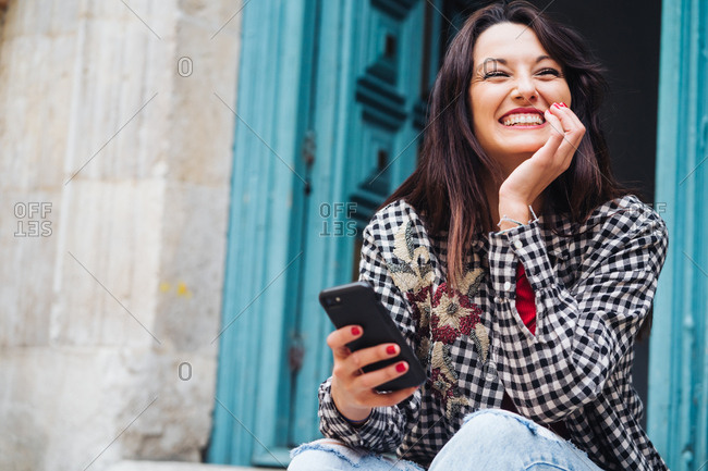 Young Brunette Woman With Red And Plaid Clothes Sitting In Front Of An Old Building With A Blueish Door While Using Her Phone.