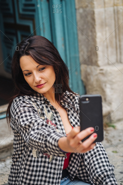 Young Brunette Woman With Red And Plaid Clothes Sitting In Front Of An Old Building With A Blueish Door While Taking A Selfie With Her Smartphone.