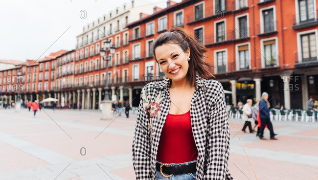 Young Brunette Woman With Red And Plaid Clothes Standing In Front Of Old Buildings In The Main Square Of A Spanish Town.