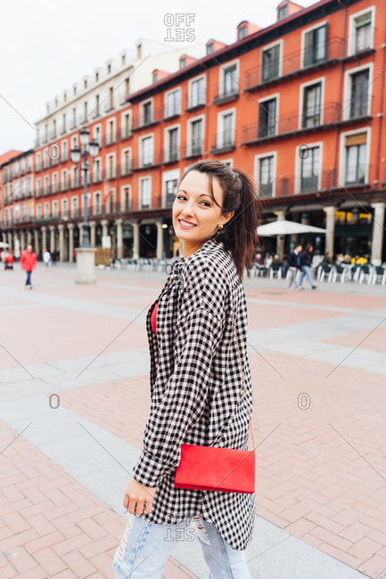 Young Brunette Woman With Red And Plaid Clothes Standing In Front Of Old Buildings In The Main Square Of A Spanish Town.