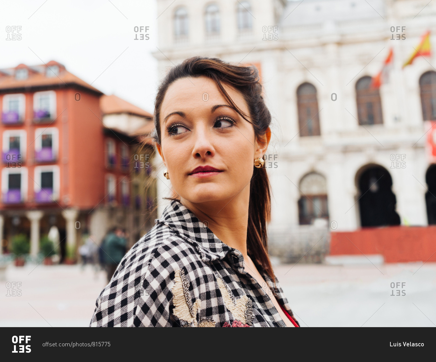 Young Brunette Woman With Red And Plaid Clothes Standing In Front Of Old Buildings In The Main Square Of A Spanish Town.