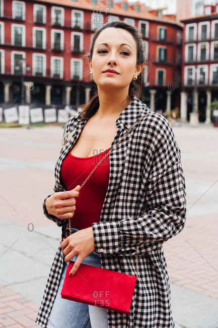 Young Brunette Woman With Red And Plaid Clothes Standing In Front Of Old Buildings In The Main Square Of A Spanish Town.