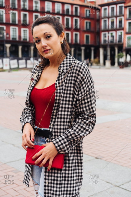 Young Brunette Woman With Red And Plaid Clothes Standing In Front Of Old Buildings In The Main Square Of A Spanish Town.
