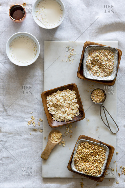 Oat grains, rolled oats, oat grist and oat milk in wooden bowls and white cups on white marble board over white background