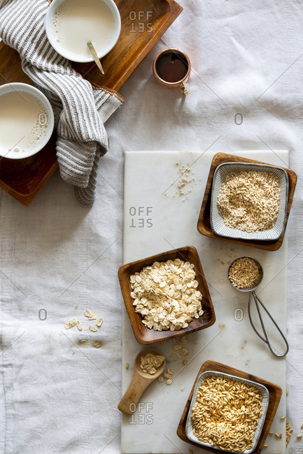 Oat grains, rolled oats, oat grist and oat milk in wooden bowls and white cups on white marble board over white background