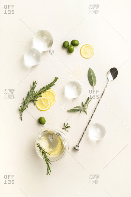 Overhead layout of minimal cocktail ingredients including large ice spheres, lemon wheels, herbs and olives on pale yellow surface.