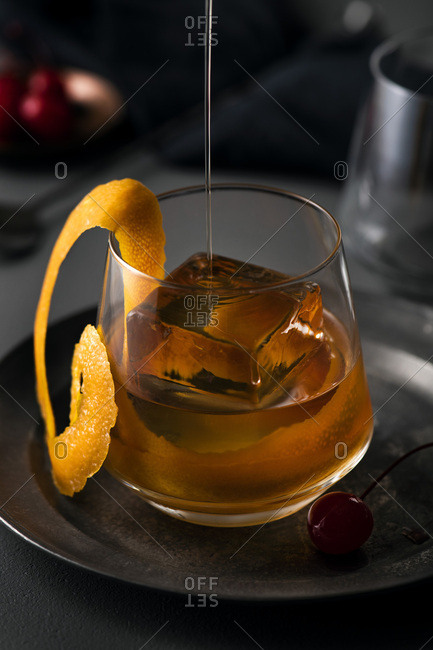 Old fashioned cocktail being poured into glass with large ice cube and orange swirl garnish.
