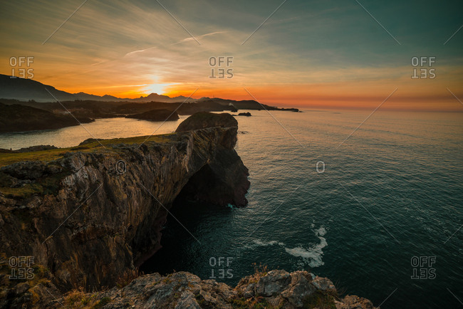 Panoramic view of huge rocky cliffs above rippled water against sunset sky, Asturias, Spain